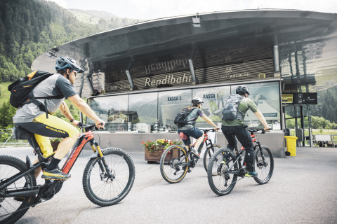 Drei Radfahrer mit Helmen fahren zu einer modernen Lodge in den Bergen, umgeben von grüner Landschaft.