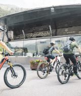 Trois cyclistes casqués et équipés de sacs à dos arrivent devant un lodge moderne dans un décor alpin.