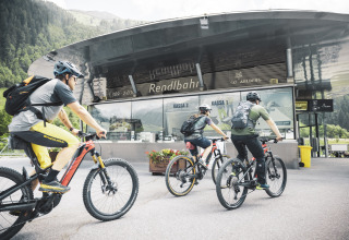 Drie fietsers met helmen en rugzakken naderen een moderne lodge in een bergachtig en groen landschap.