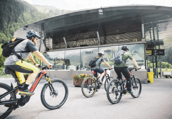 Tres ciclistas con cascos y mochilas llegan a una moderna cabaña rodeada de montañas y vegetación.