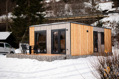 Een modern, met hout bekleed lodgehuis met terras en stoelen, omringd door sneeuw en bomen.