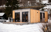 Un lodge moderne en bois avec terrasse et chaises, entouré de neige et d’arbres en hiver.