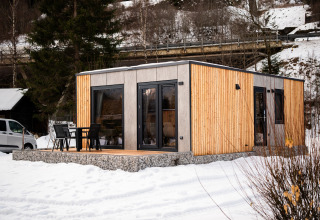 Een modern, met hout bekleed lodgehuis met terras en stoelen, omringd door sneeuw en bomen.