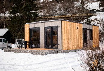 Un lodge moderne en bois avec terrasse et chaises, entouré de neige et d’arbres en hiver.
