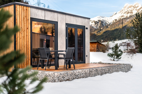 Et moderne træ- og betonlodge med terrasse og stole, omgivet af sne og bjerge i baggrunden.
