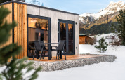 Un lodge moderne en bois et béton avec terrasse et chaises, entouré de neige et de montagnes.