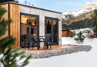 Et moderne træ- og betonlodge med terrasse og stole, omgivet af sne og bjerge i baggrunden.