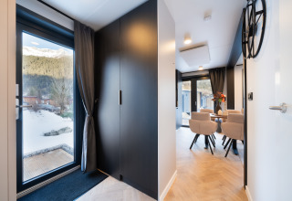 Modern entré og spisestue i en hytte med lyse møbler og store vinduer, udsigt til snedækkede bjerge.
