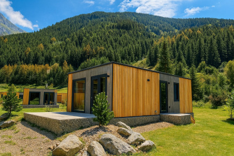 Moderne houten lodge in de bergen, omgeven door groene bomen en gras onder een heldere blauwe lucht.