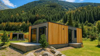 Modern wooden lodge in a mountain setting, surrounded by lush trees and grass under a bright blue sky.