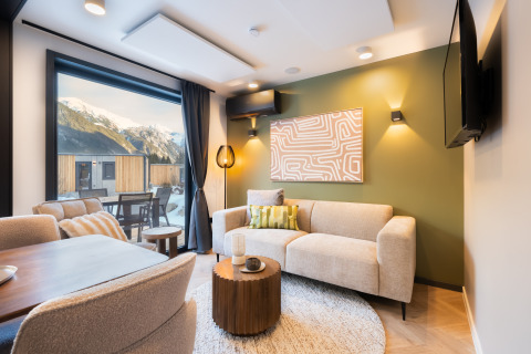 Moderne lodge woonkamer met grote ramen, bergzicht, beige bank en groene accentmuur bij zonsondergang.
