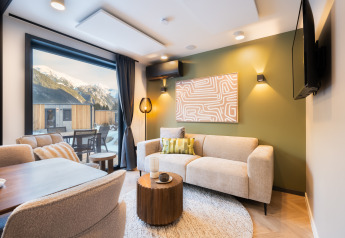 Salon de lodge moderne avec grandes fenêtres, vue sur la montagne, canapé beige et mur d’accent vert.