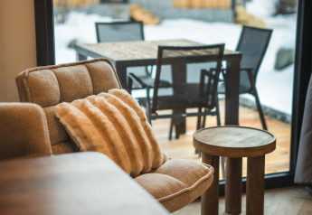 Acogedora sala de estar con sillón y mesa, vista a la terraza nevada de una cabaña de montaña a través de la ventana.
