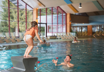 Un enfant s'apprête à sauter dans une piscine intérieure où un adulte l'attend dans l'eau d'un lodge.