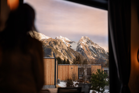 Een persoon kijkt vanuit een lodge-raam naar besneeuwde bergen en moderne houten huisjes op de voorgrond.