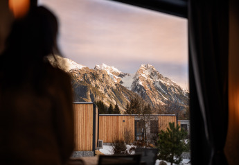 Une personne regarde par la fenêtre d’un lodge vers des montagnes enneigées et des chalets en bois modernes.