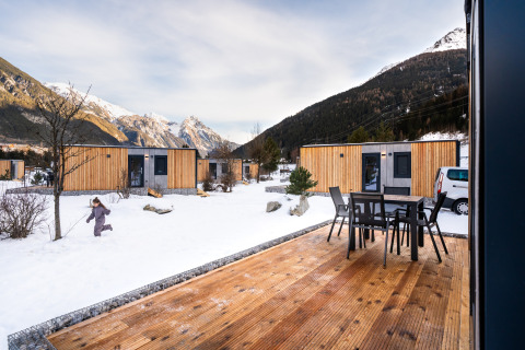 Een lodge met houten huisjes in de sneeuw, bergzicht, buitenterras en een kind dat buiten speelt.