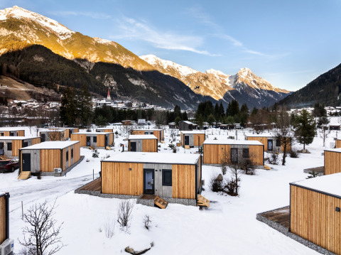 Houten lodges in een besneeuwd landschap, omgeven door bergen en een dorp onder een heldere lucht.