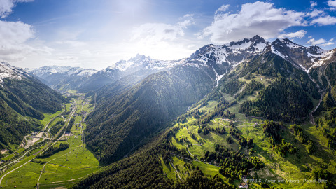 Panoramic view of a lush valley and snow-capped mountains, featuring a lodge under a sunny blue sky.