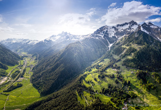 Panoramisch zicht op een groene vallei en besneeuwde bergen, een lodge onder een stralende blauwe hemel.