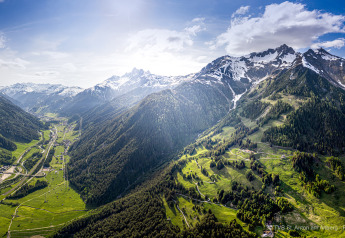 Vista panorámica de un verde valle y montañas nevadas, destacando una cabaña bajo un cielo soleado.