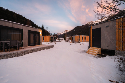 Sneklædte træhytter ved en lodge omgivet af bjerge, med lyserøde skyer på himlen ved solnedgang.