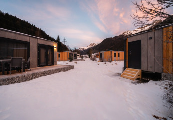 Cabañas modernas y lodges cubiertos de nieve entre montañas, cielo al atardecer con nubes rosadas.