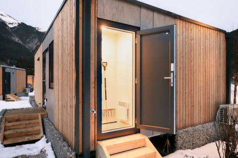 Indgang til en moderne træhytte med åben dør, udsigt til sneklædte bjerge i baggrunden ved en lodge.