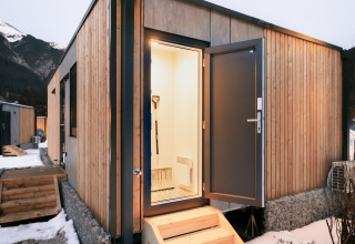 Indgang til en moderne træhytte med åben dør, udsigt til sneklædte bjerge i baggrunden ved en lodge.