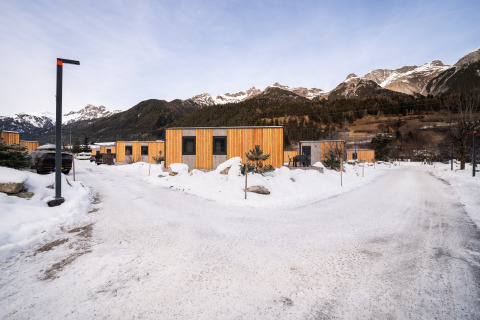 Moderne houten lodges op een besneeuwd vakantiepark met bergen op de achtergrond en brede paden.