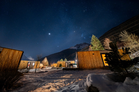 Stjerneklar nat over en træhytte-lodge i snemiljø med bjerge i baggrunden og udendørs belysning.