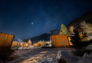 Stjerneklar nat over en træhytte-lodge i snemiljø med bjerge i baggrunden og udendørs belysning.