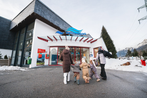 Gezin in winterkleding arriveert bij Wellnesspark lodge, omringd door besneeuwde bergen en dennen.