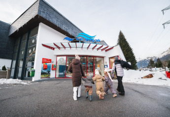 Famille en vêtements d'hiver arrivant au lodge Wellnesspark, entouré de montagnes enneigées et de sapins.