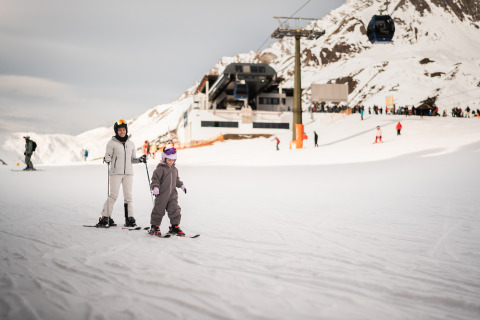 En voksen og et barn står på ski foran en bjerglodge omgivet af sne og andre skiløbere.