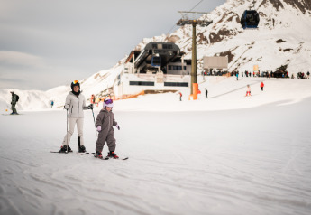 Un adulto y un niño esquiando juntos cerca de un refugio de montaña, rodeados de nieve y esquiadores.