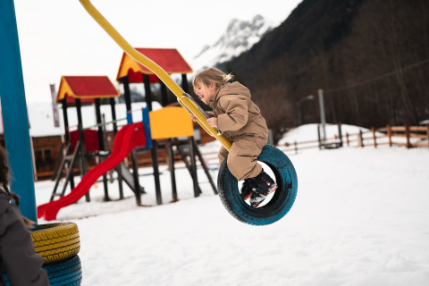 Child in brown snowsuit swings on a tire swing at a snowy playground near a mountain lodge.