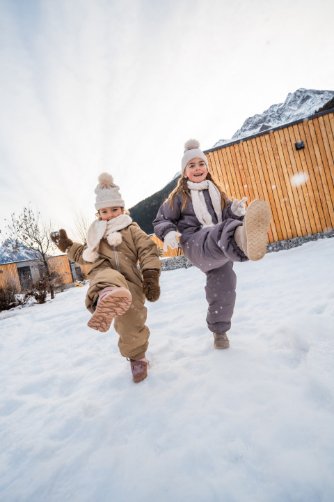 Twee kinderen spelen vrolijk in de sneeuw bij een houten lodge, warm gekleed in winteroutfits.