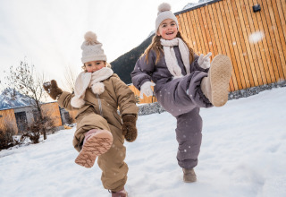 Twee kinderen spelen vrolijk in de sneeuw bij een houten lodge, warm gekleed in winteroutfits.