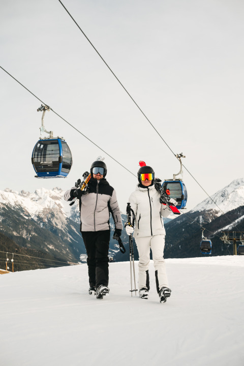 Twee skiërs in winterkleding lopen met ski’s en stokken bij een kabelbaan in een besneeuwd skigebied.
