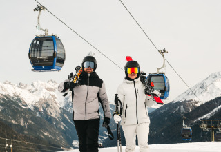 Twee skiërs in winterkleding lopen met ski’s en stokken bij een kabelbaan in een besneeuwd skigebied.