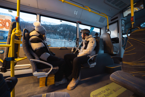 Twee mensen in winterjassen zitten tegenover elkaar in een bus, besneeuwde bergen buiten het raam.