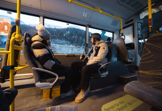 Twee mensen in winterjassen zitten tegenover elkaar in een bus, besneeuwde bergen buiten het raam.