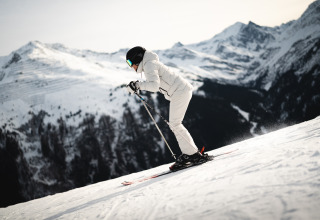Skiër in witte uitrusting skiet van een besneeuwde helling met bergachtig landschap op de achtergrond.