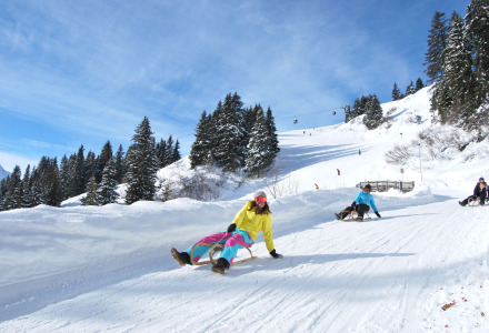 Des personnes font de la luge sur une piste enneigée bordée d’arbres et de montagnes sous un ciel bleu.
