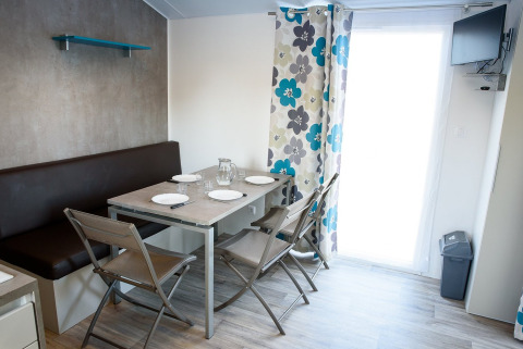 Coin repas moderne dans un mobil-home avec table dressée, chaises, banquette et rideaux à fleurs.