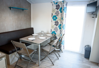 Coin repas moderne dans un mobil-home avec table dressée, chaises, banquette et rideaux à fleurs.