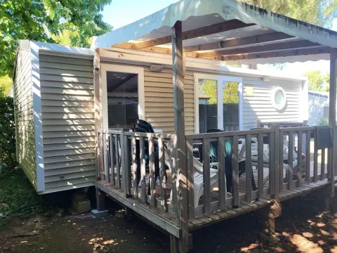 Photo d’un mobil-home avec terrasse en bois couverte et chaises, entouré de verdure et d’arbres.
