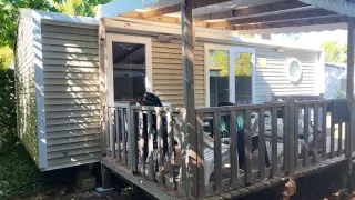 Foto di una casa mobile con veranda in legno coperta e sedie da giardino, circondata dal verde.