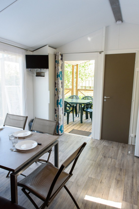 Interior de una casa móvil con mesa de comedor, sillas y puerta abierta hacia una terraza cubierta con asientos.